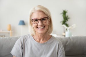 Smiling middle aged mature grey haired woman looking at camera, happy old lady in glasses posing at home indoor, positive single senior retired female sitting on sofa in living room headshot portrait